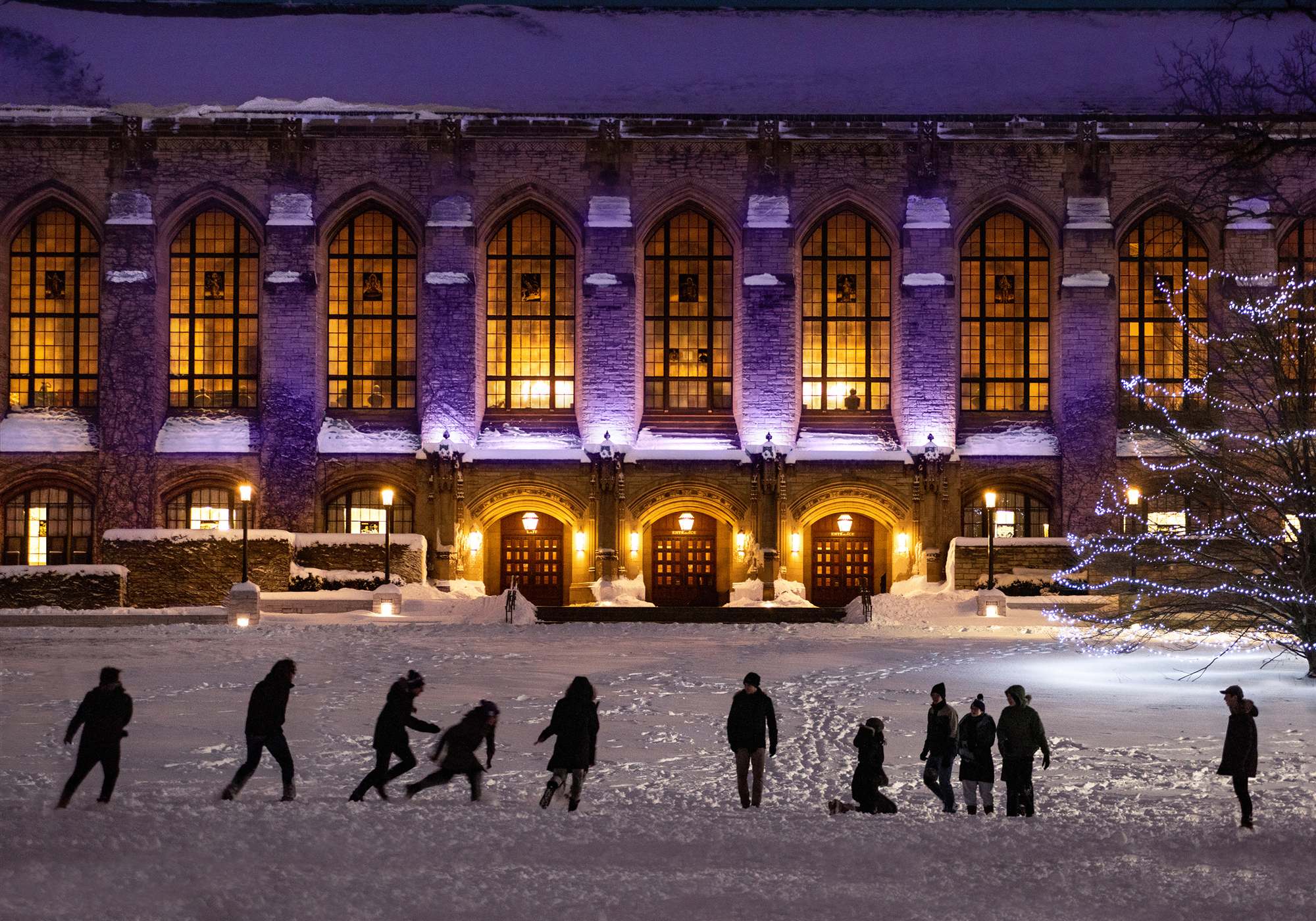 Deering Library lit purple on a winter night, with people walking across the snowy lawn